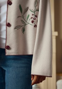 Šviesiai smėlinis kardiganas su raudonais mygtukais ir išsiuvinėtoms žalioms lapams su raudonomis uogomis, turi švelnią tekstūrą ir kontrastingą burgundiško atspalvio siūlą.