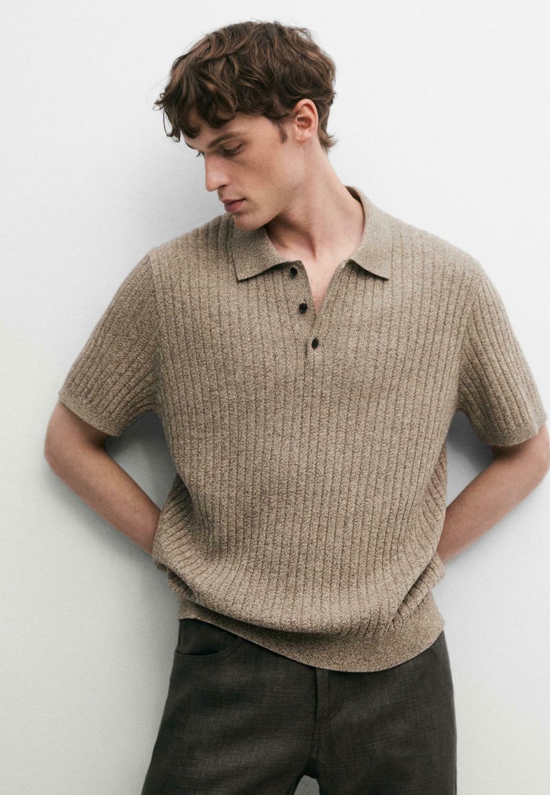 Young man wearing a beige ribbed short-sleeve polo shirt and dark pants, standing with hands behind back, looking downward to side.