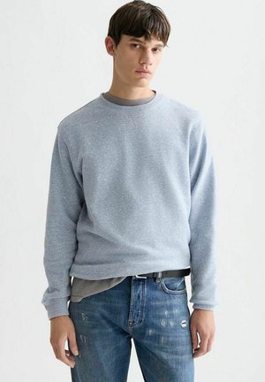 Junger Mann mit kurzen braunen Haaren, der einen hellgrauen Pullover über einem grauen T-Shirt und blaue zerrissene Jeans vor einem schlichten weißen Hintergrund trägt.
