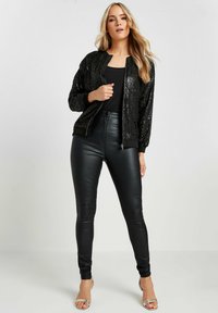 Giacca bomber nera con paillettes, polsini a costine e chiusura con zip, abbinata a leggings neri in ecopelle a vita alta e tacchi argentati con cinturini.