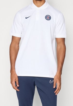 Homme portant un polo blanc Paris Saint-Germain avec un logo Nike bleu marine, associé à un pantalon Nike bleu marine, debout devant un fond uni.