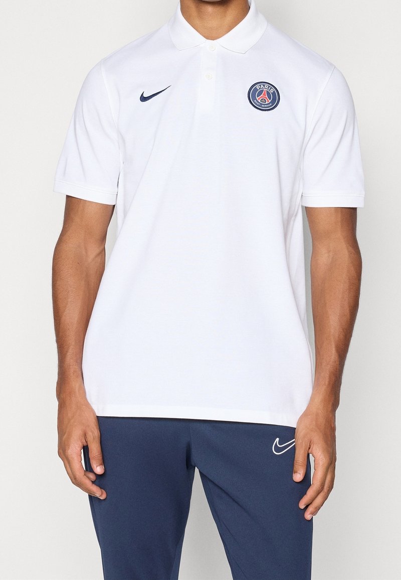Homme portant un polo blanc Paris Saint-Germain avec un logo Nike bleu marine, associé à un pantalon Nike bleu marine, debout devant un fond uni.
