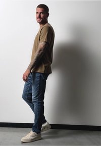 Hnědé tričko s krátkým rukávem, modré slim-fit džíny a krémové tenisky. Tetování na pažích viditelné; stojící na neutrálním pozadí.