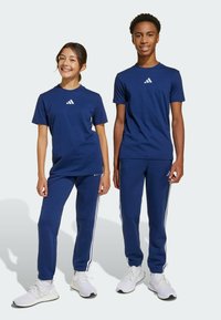T-shirts de sport bleu marine et pantalon de survêtement avec des accents blancs à trois bandes, fabriqués en tissu doux, associés à des baskets blanches, présentés par deux jeunes.