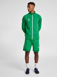 Grøn atletisk tracksuit med en jakke med lynlås og shorts, der har strukturerede paneler og sølvdetaljer på shortsene. Tilbehøret inkluderer hvide sokker og blå sneakers.