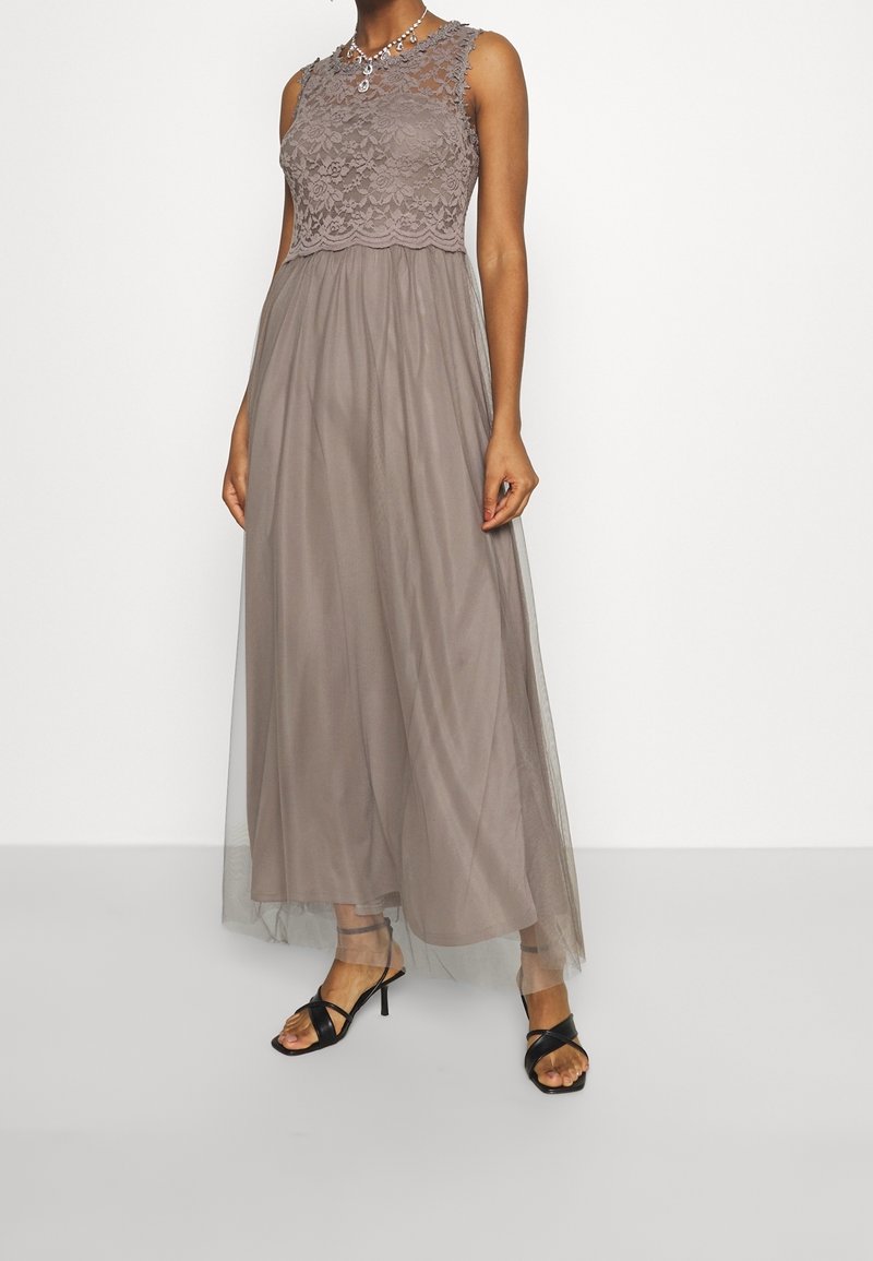 Femme portant une robe maxi taupe sans manches en dentelle et tulle, des talons noirs à brides et un collier argenté avec des pendentifs.