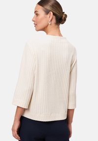 Femme avec les cheveux en chignon portant des boucles d'oreilles en argent, un pull en maille texturée couleur off-white avec des manches 3/4, et un pantalon noir, de profil devant un fond blanc.