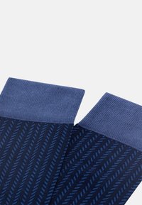 Tumšu zilo zeķu pāris ar gaiši zilu diagonālu svītru rakstu un vienkrāsainiem gaiši ziliem maliem, izstādīts uz balta fona.