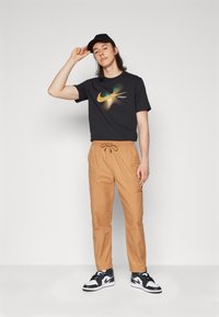 Crni Nike majica s grafikom u zelenoj i žutoj boji, bež hlače s vezicom, i crno-bijele tenisice. U stojećem položaju, nosi crnu kapu.