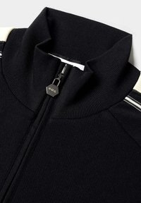 Svart zip-up jacka med hög krage, ribbad textur och vita ränder på axlarna, med en metallisk dragkedjehållare.