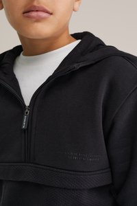 Svart zip-up hoodie med en texturerad bröstpanel, huva och vit inuti tröjan som syns. Har logotyp på dragkedjan och tryckdetalj på tyget.