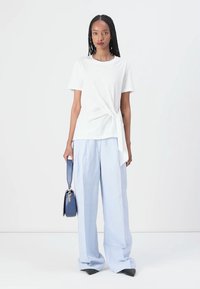 Tricou alb cu nod, cu mâneci scurte, asortat cu pantaloni largi de culoare albastru deschis. Modelul ține o geantă mică albastră și poartă pantofi negri cu vârful ascuțit.