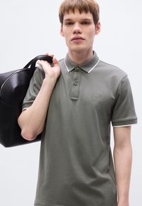 Γκρι πόλο πουκάμισο με λευκό περίγραμμα, δύο κουμπιά και κηλίδα με λογότυπο στο στήθος. Το μοντέλο κρατάει μια μαύρη τσάντα πάνω σε έναν ώμο μπροστά από ένα απλό φόντο.