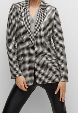 Personne portant un blazer pied-de-poule noir et blanc sur un haut gris boutonné, associé à un pantalon en cuir noir sur un fond uni.
