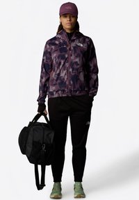 Violeta maskēšanas jaka ar oranžiem akcentiem, melnas bikses, zaļi apavi un melna soma. Cepure atbilst jaciņas krāsai un ir ar logotipu.