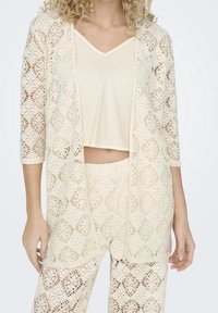 Lace cardigan in off-white with three-quarter sleeves, open front, and detailed cut-out patterns, worn over a matching cropped top and pants.