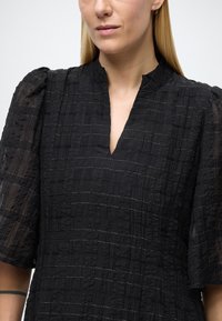 Juoda suknelė iš tekstūruotos audinio su horizontaliomis juostelėmis, su gilia V formos iškirpte ir trumpomis pūstomis rankovėmis, pademonstruojanti sudėtingus detalių elementus.