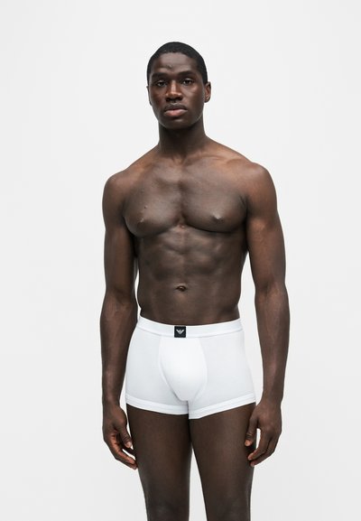 Homme musclé à la peau foncée, debout face à l’avant, portant un boxer blanc avec un logo noir sur la ceinture, sur un fond clair uni.
