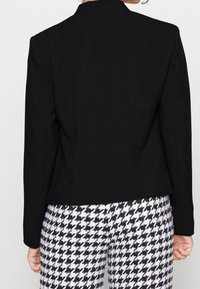 Blazer noir sur mesure avec une coupe structurée, doté d'un col cranté discret, de manches longues et d'un tissu lisse. Associé à un pantalon à motif pied-de-poule.