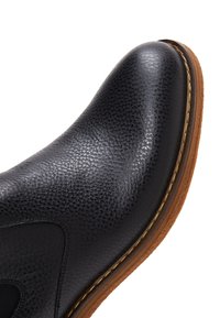 Bottine en cuir noir avec surface texturée, forme courbée, semelle en caoutchouc marron clair et couture contrastante le long du bord.
