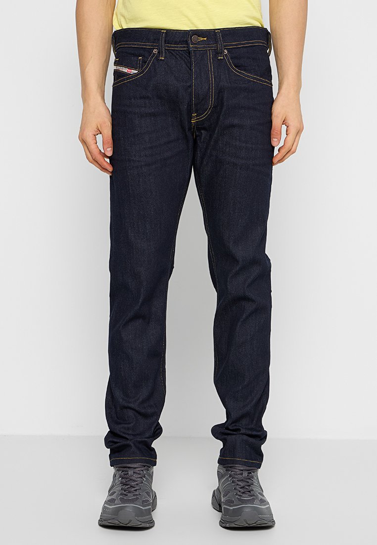 Dunkelblaue Jeans mit gelben Nähten, geradem Schnitt, vorderen Taschen und einem kleinen Etikett an einer Tasche. Getragen mit grauen Schuhen.