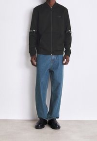 Giacca nera con zip e colletto alto, dotata di strisce riflettenti sulle maniche e due tasche frontali, abbinata a jeans in denim blu sbiadito.