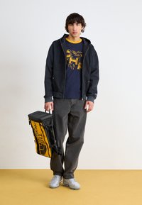 Schwarze Zip-Hoodie-Jacke, marineblaues Grafik-T-Shirt, graue Hose, trägt einen gelb-schwarzen Rucksack mit Logo, graue Sneakers.