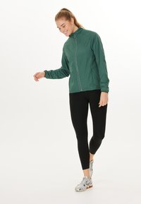 Lichte groene jack met een voorrits, elastische cuffs en witte pipingdetails, gecombineerd met zwarte leggings en gemusterde hardloopschoenen.