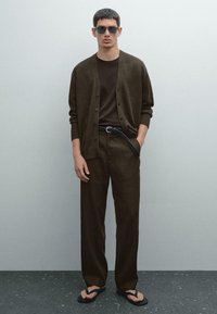 Brown knitted cardigan over a brown t-shirt, paired with matching loose pants. Black belt with braided detail, black flip-flops, dark sunglasses.