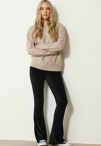Donna con lunghi capelli biondi che indossa un maglione beige a trama a treccia, pantaloni neri a zampa d'elefante e sneaker bianco-neri, in piedi contro un muro bianco.