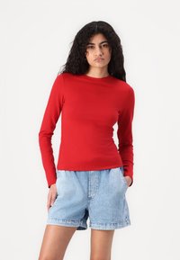 Tricou roșu cu mânecă lungă și guler rotund, asortat cu șorturi din denim ușor albastru. Materialul pare neted cu o siluetă ajustată.