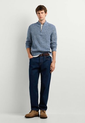 Jeune homme debout portant un pull bleu à demi-fermeture éclair, un jean bleu foncé, une ceinture marron et des bottes à lacets beige, sur un fond clair uni.