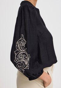 Blusa nera con maniche a sbuffo, caratterizzata da un ricamo dettagliato bianco sulla manica sinistra e polsini elastici. Tessuto leggero.