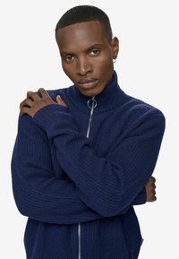 Laivastonsininen vetoketjullinen neulepaita, jossa on ribbikuvio, korkea kaulus ja hopeanväriset yksityiskohdat. Paita on pitkähihainen ja istuu tiiviisti.