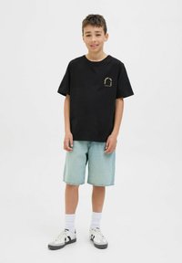 Camiseta de algodón negra con cuello redondo, que presenta un pequeño gráfico amarillo. Combinada con pantalones cortos de mezclilla claros y calcetines blancos con zapatillas negras.