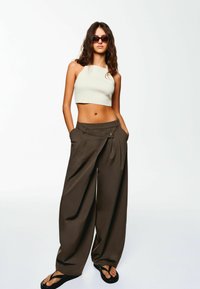 Top beige accorciato abbinato a pantaloni larghi marrone scuro con pieghe e dettagli con bottone, completato da flip-flop neri.