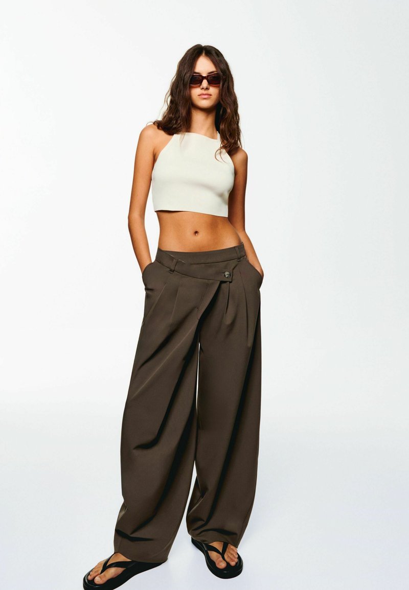 Top beige accorciato abbinato a pantaloni larghi marrone scuro con pieghe e dettagli con bottone, completato da flip-flop neri.