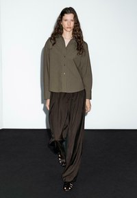 Olive green button-up shirt paired with loose, dark brown pants. Model wears black strappy sandals. Clean, minimalist background.