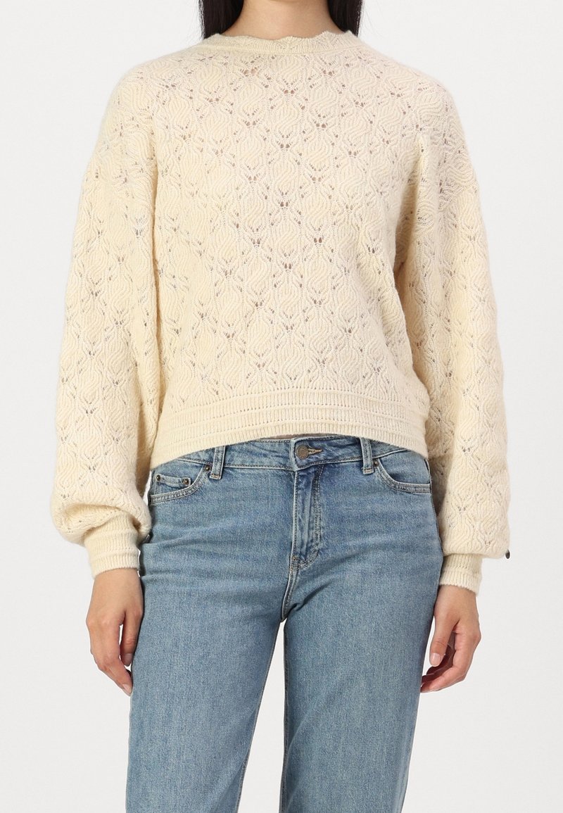 Femme portant un pull en maille crème à manches longues avec un motif texturé, associé à un jean bleu clair taille haute.