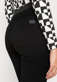 Calças jeans pretas com um design justo, apresentando uma cintura média, bolsos traseiros e tecido texturizado. Etiqueta visível acima do bolso direito.