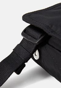 Svart messenger bag med teksturert stoff, justerbar stropp og plastlås. Har en metalltrykkdetalj på siden.