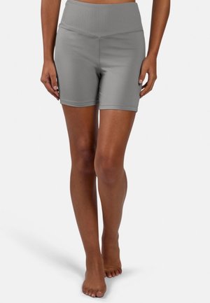 Personne portant un short de sport gris à taille haute avec une ceinture côtelée, debout pieds nus sur un fond blanc.