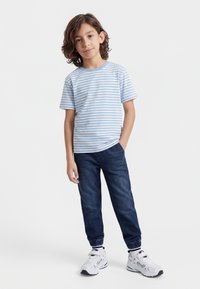 Keskmise pikkusega pruunide juustega poiss kannab helesinist ja valget triibulist t-särki, tumedate siniste dressipükstega ja valgete tossudega, seistes ühe käega püksitaskus.