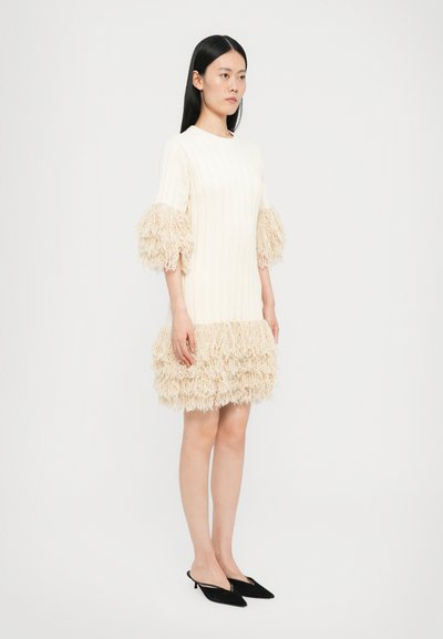 Robe en tricot crème avec une texture côtelée, des manches bouffantes à franges et un ourlet volanté. Assortie à des escarpins noirs à bride arrière.