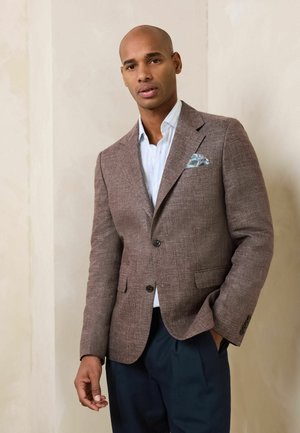Mann trägt braunen strukturierten Blazer, hellblaues Hemd, marineblaue Hose und gemustertes Einstecktuch, steht vor einer beigen Wand.