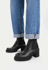 Czarne skórzane botki za kostkę z masywną, teksturowaną podeszwą, kwadratowym noskiem i elastycznymi panelami po bokach, zestawione z podwiniętymi niebieskimi dżinsami.