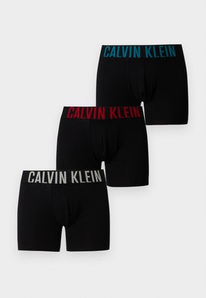 Młody mężczyzna w czarnych bokserkach Calvin Klein stoi boso na wykładzinie, pozując obok ciemnej skórzanej sofy na tle gładkiej białej ściany.