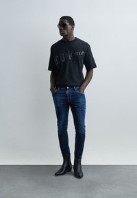 Homme portant un t-shirt noir avec motif graphique, un jean slim bleu foncé, des bottines noires et des lunettes de soleil, debout les mains dans les poches contre un mur gris.