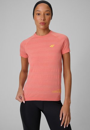 Kobieta ubrana w koralowy sportowy t-shirt z krótkim rękawem z żółtym logo i czarne legginsy, stoi na jednolitym szarym tle.