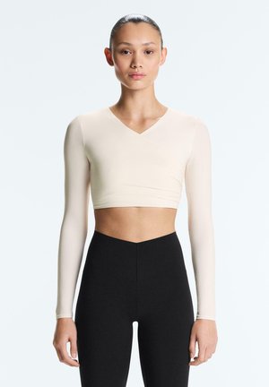 Vrouw met haar naar achteren getrokken draagt een beige cropped top met lange mouwen en een zwarte broek met hoge taille, staand tegen een effen achtergrond.